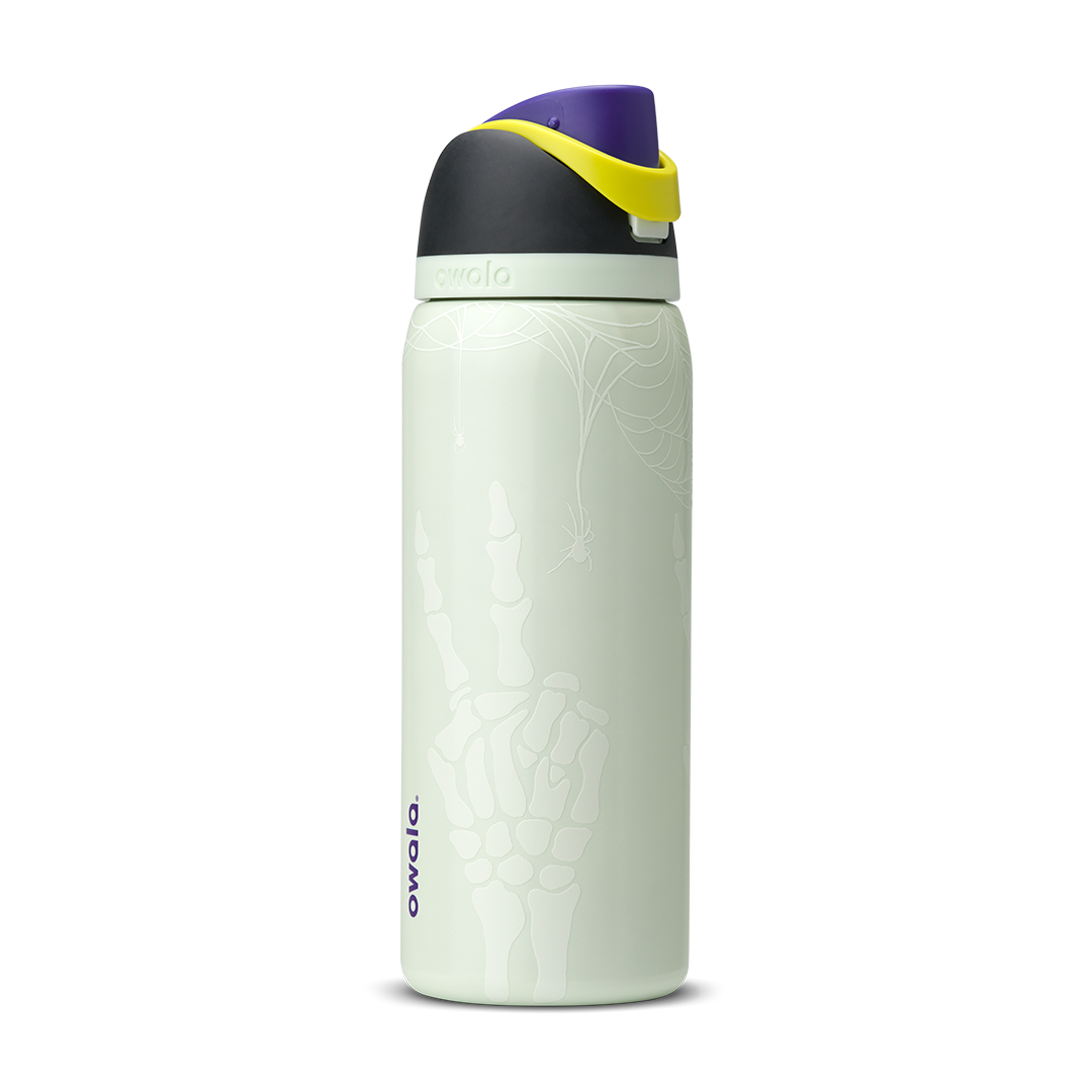 Owala FreeSip 32oz in RIP, pale mint green insulated stainless steel bottle with white skeleton-hand pattern body, dark charcoal lid, yellow carry loop, purple flip cap, and pale green accent ring; glows in the dark